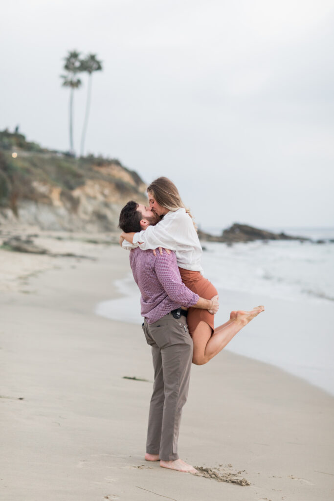 A happy couple standing on the beach, the man is picking the woman up and they are kissing.