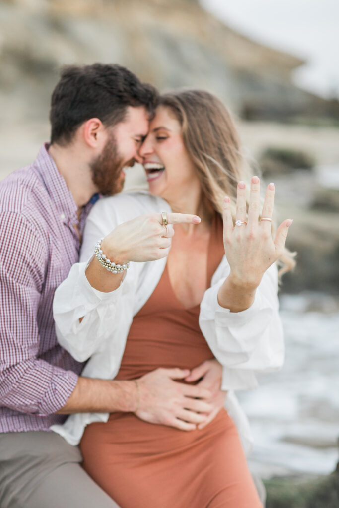 A newly engaged couple snuggling on the rocks in Laguna Beach, CA. The woman is smiling and laughing while pointing at the engagement ring on her finger.