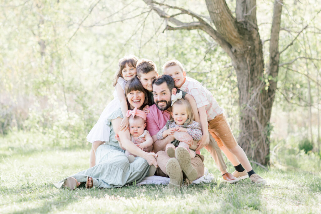 A family with a mother, father, and five small children are piled together in a green, wooded area, filled with light.