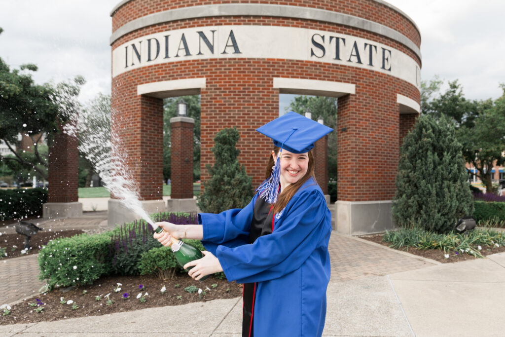 A college graduate celebrating by popping a bottle of champagne in front of their university's sign.