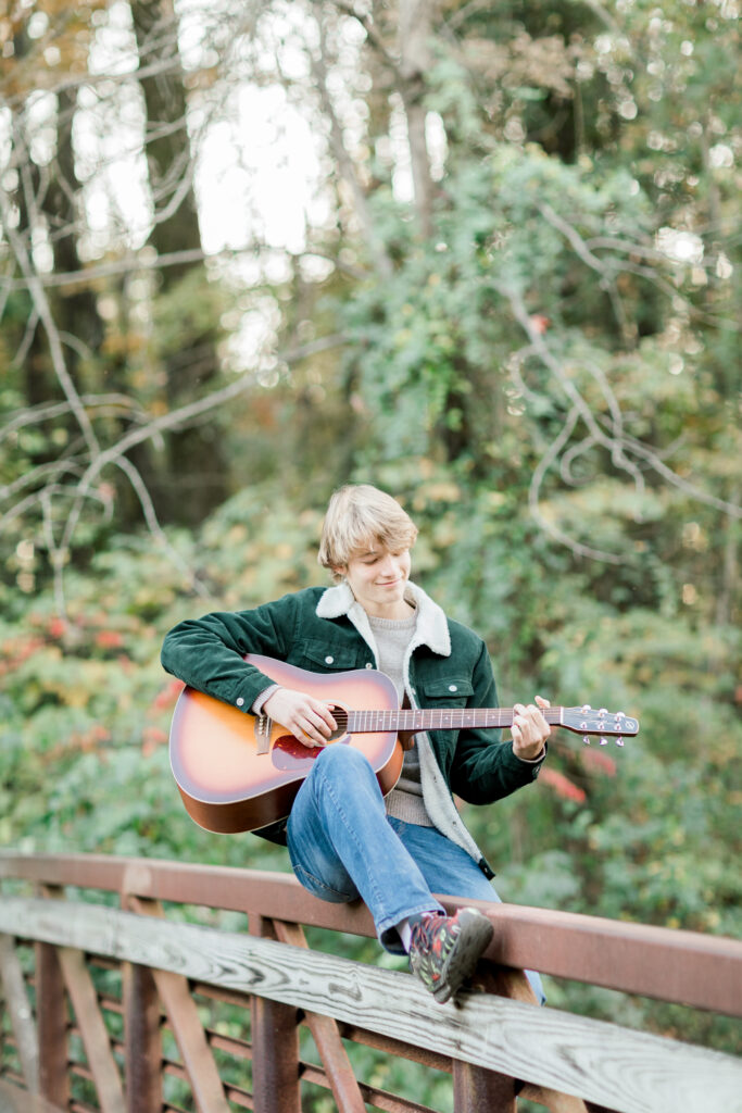 A high school senior boy sitting on the railing of a bridge playing an acoustic guitar.