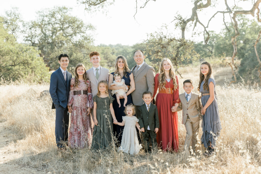 A large family consisting of a mother, father, and 10 children dressed in their Sunday best, gathered together in a field of tall, golden grass.