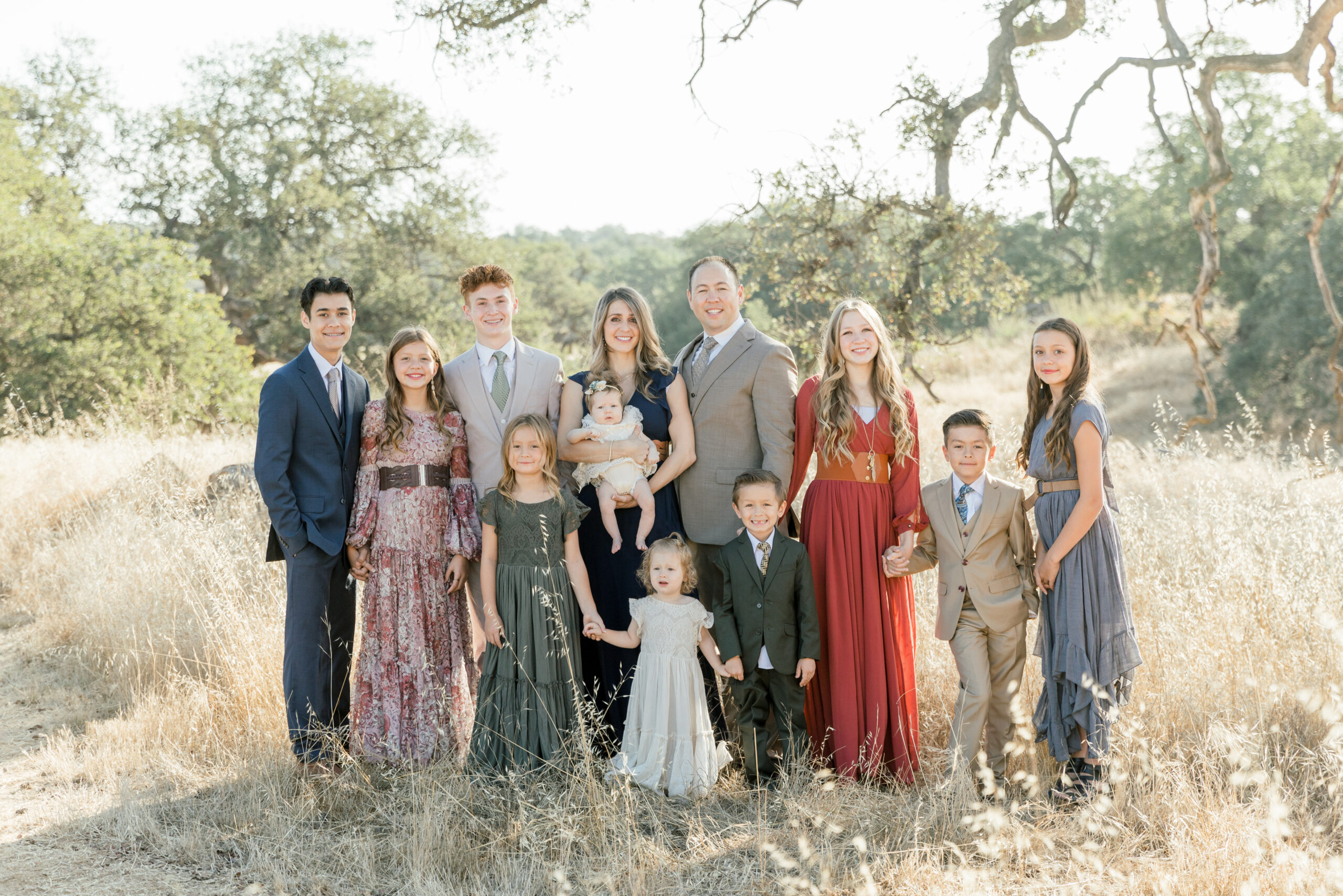 A large family consisting of a mother, father, and 10 children dressed in their Sunday best, gathered together in a field of tall, golden grass.