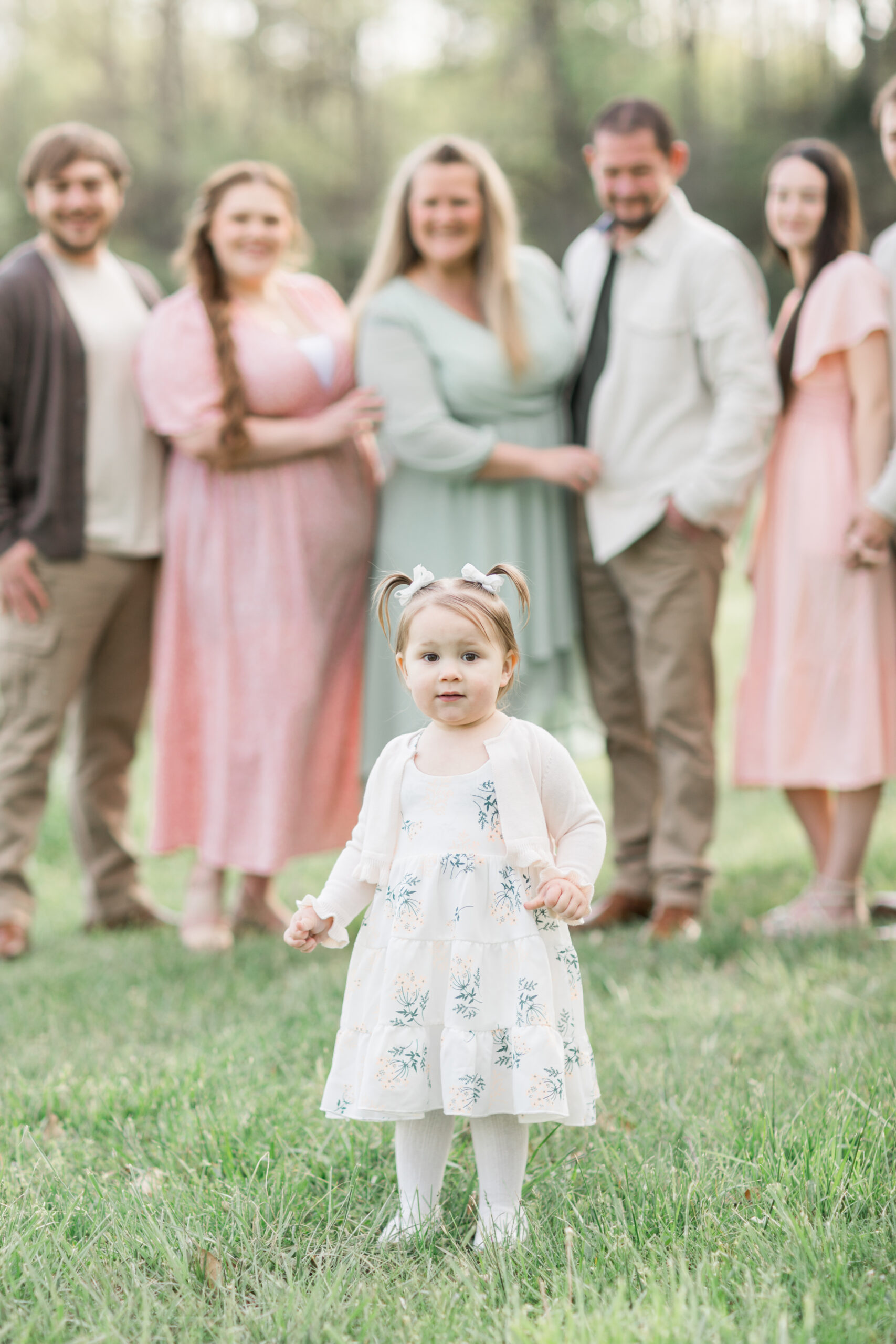 A toddler girl in a pretty white, floral print dress standing just in front of her extended family, who are in the background smiling at her.