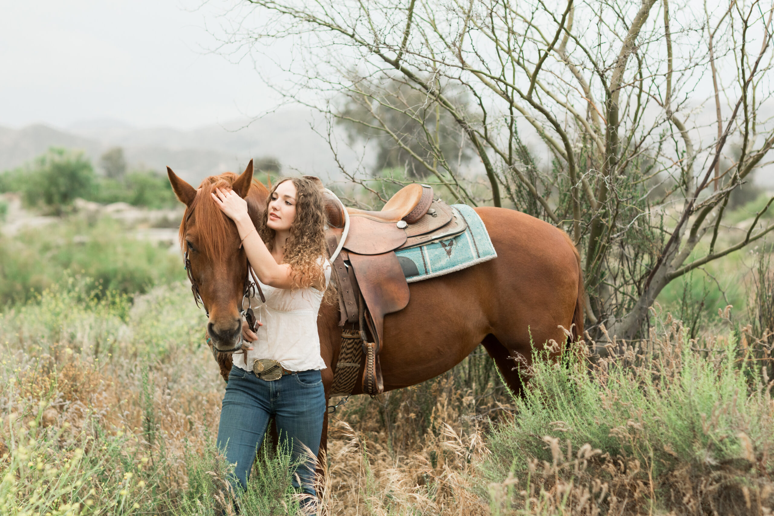 A beautiful high school graduate girl standing next to a chestnut horse tacked in turquoise western saddlery and standing in a field of tall grass and sagebrush.
