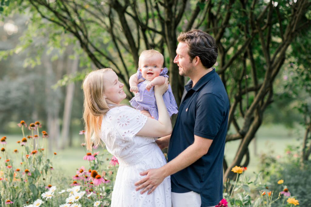 An adorable family with a pregnant mother in a white dress holding her toddler daughter and her husband lovingly looking toward them both.