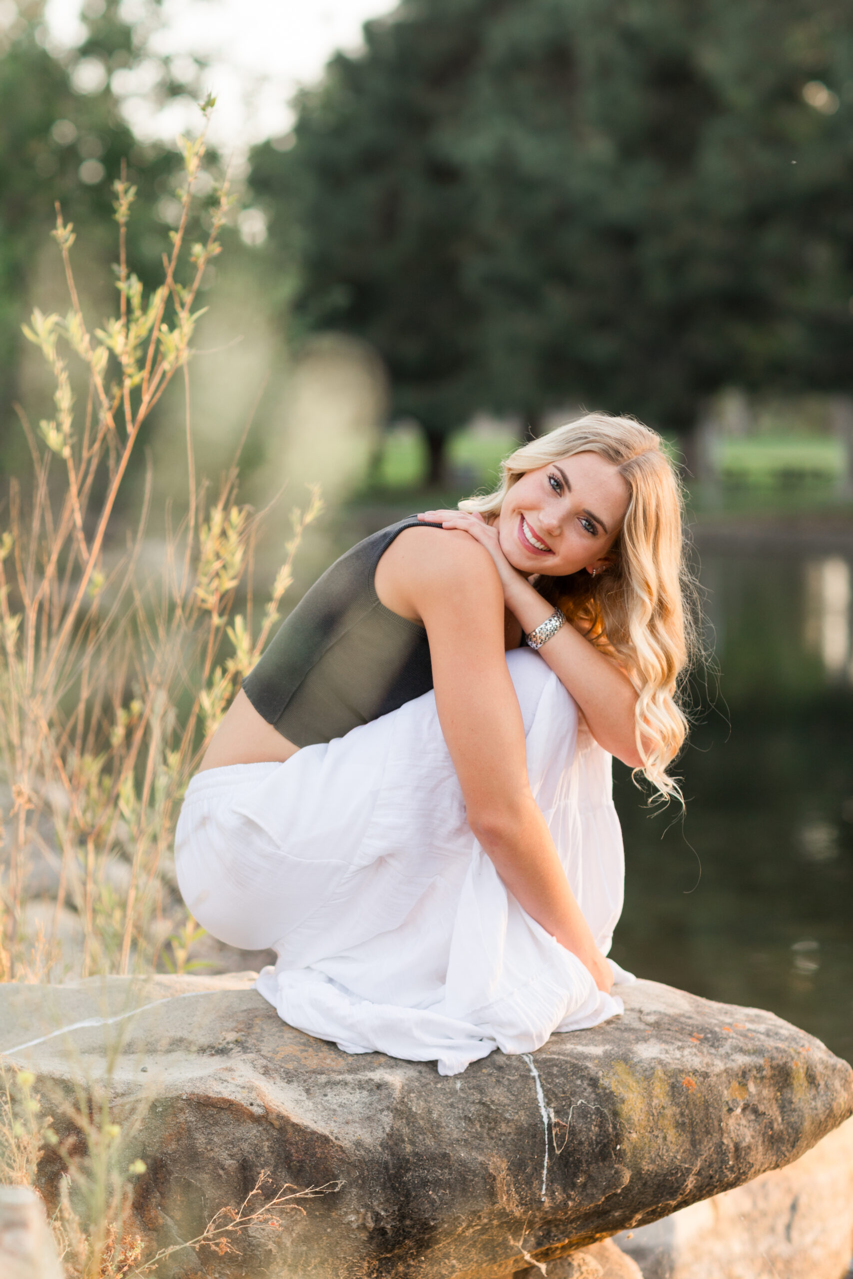 a high school senior girl kneeling on a rock next to a pond, framed by tall golden grass and golden light.