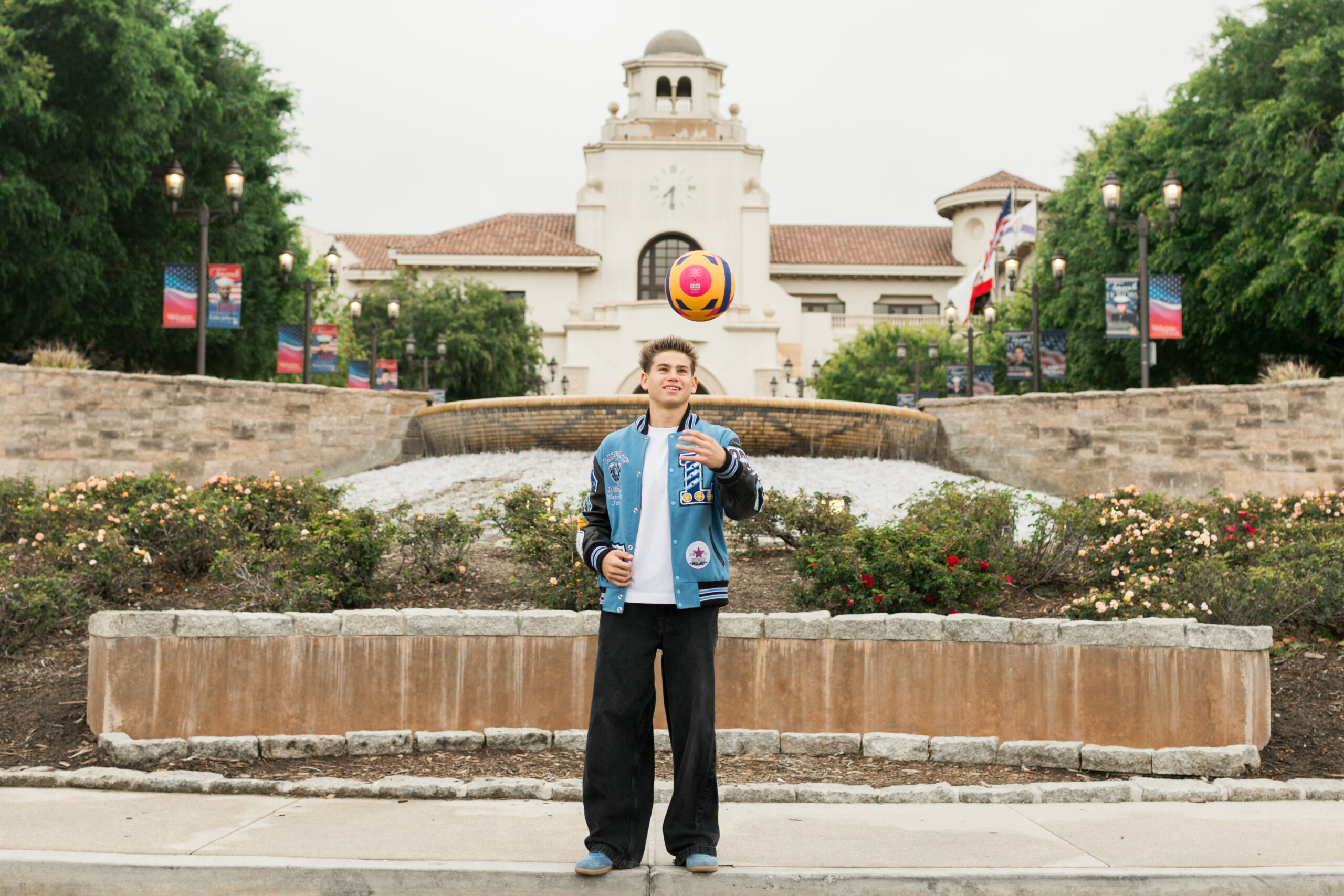 A High School Senior boy standing in front of a large building with a water fountain, tossing a water polo ball in the air.